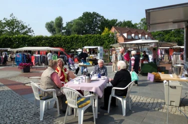 Menschen sitzen bei Kaffee an Tischen, umgeben von Marktständen mit Kleidung und regionalen Produkten.
