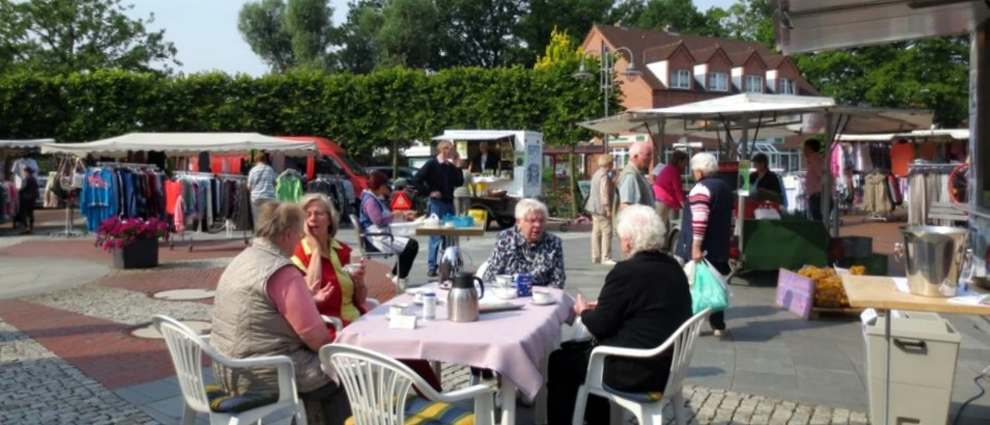 Menschen sitzen bei Kaffee an Tischen, umgeben von Marktständen mit Kleidung und regionalen Produkten.