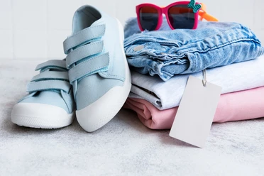 Kinderflohmarkt.png Stapel Kinderkleidung, oben drauf liegt eine Sonnenbrille und Kinderschuhe sind an den Stapel angelehnt