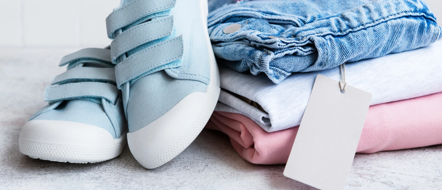 Kinderflohmarkt.png Stapel Kinderkleidung, oben drauf liegt eine Sonnenbrille und Kinderschuhe sind an den Stapel angelehnt