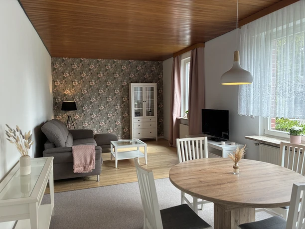Heller Wohnraum mit Essplatz, grauem Sofa, Holzdecke und floraler Tapete an der Rückwand.