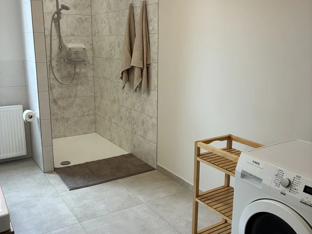 Helles Bad mit bodengleicher Dusche, beige gemusterten Wandfliesen und beigen Handtüchern. Rechts steht eine weiße Waschmaschine neben einem kleinen Holzregal, auf dem Boden liegt eine braune Matte vor der Dusche. Der Raum wirkt modern, praktisch und großzügig gestaltet.
