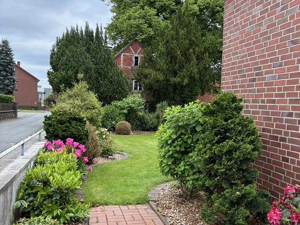 Roter Klinkerweg führt durch einen gepflegten Garten mit grüner Rasenfläche, bunten Blüten und dichten Sträuchern. Im Hintergrund stehen hohe Bäume und ein rotes Backsteinhaus unter bewölktem Himmel, umgeben von weiteren Wohnhäusern am ruhigen Straßenrand.