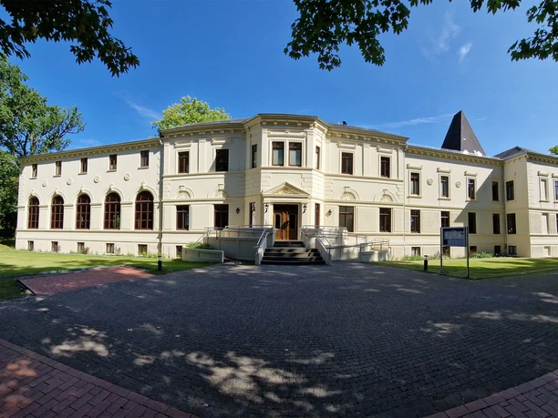 Altes-Kurhaus-Panorama.jpg Altes Kurhaus mit großen Fenstern, umgeben von Bäumen, in Bad Zwischenahn unter blauem Himmel.