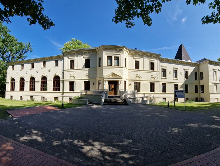 Altes-Kurhaus-Panorama.jpg Altes Kurhaus mit großen Fenstern, umgeben von Bäumen, in Bad Zwischenahn unter blauem Himmel.