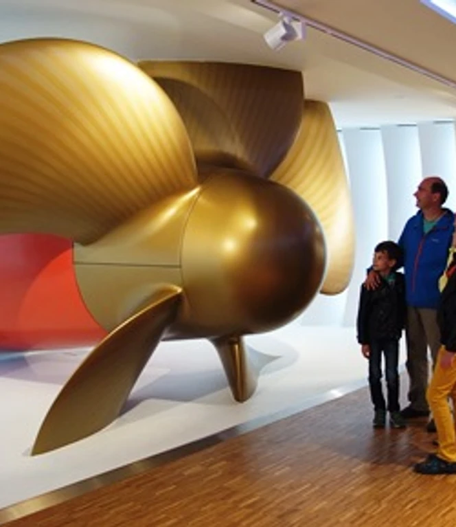 Goldene Turbine Besucherzentrum Meyerwerft.JPG Gruppe betrachtet eine große goldene Schiffsturbine im modernen Besucherzentrum der Meyer Werft.