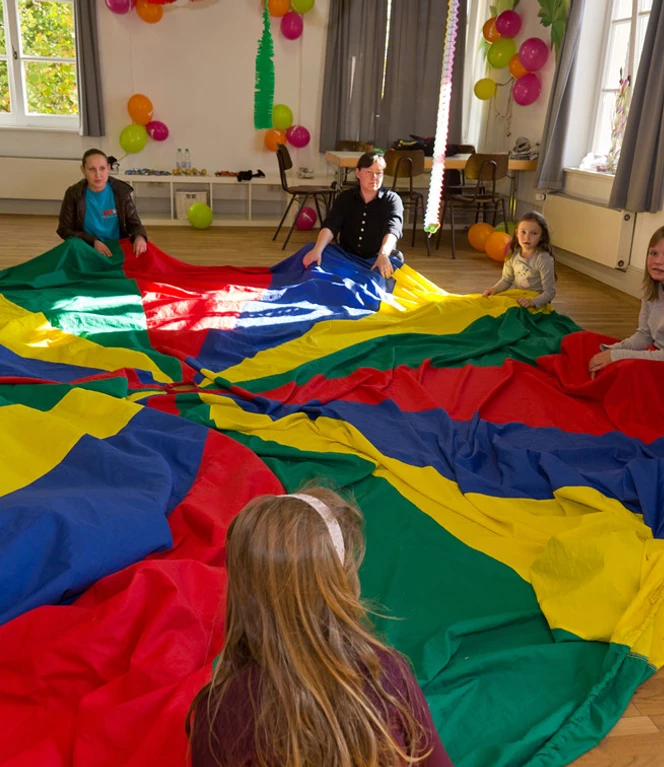 Erlebnishaus im TPZ Lingen Kinder und Erwachsene sitzen in einem Raum im Kreis und halten gemeinsam ein buntes Schwungtuch.