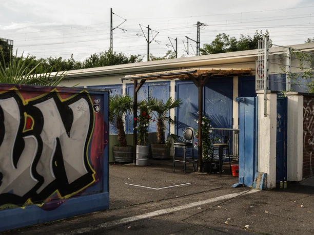 Garagen Club Außenansicht eines Club-Eingangs mit Wandgraffitis, mehreren Pflanzenkübeln und blauer Fassade.