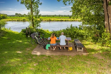 Radeln auf Emsland-Route mit Rastplatz an Ems, Emen ©Emsland Tourismus GmbH (3) Zwei Radfahrende sitzen an einem Picknicktisch am Flussufer der Ems, neben ihnen stehen Fahrräder.