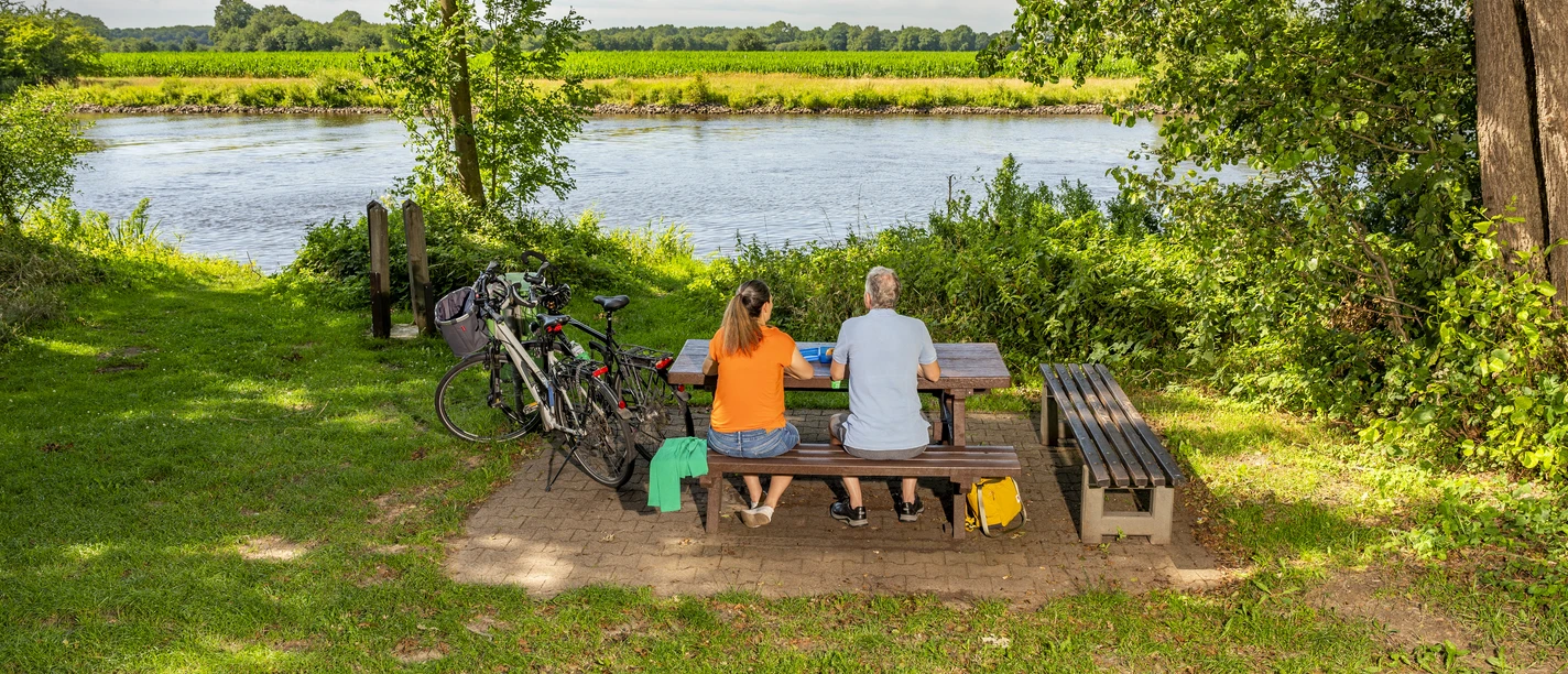 Radeln auf Emsland-Route mit Rastplatz an Ems, Emen ©Emsland Tourismus GmbH (3) Zwei Radfahrende sitzen an einem Picknicktisch am Flussufer der Ems, neben ihnen stehen Fahrräder.