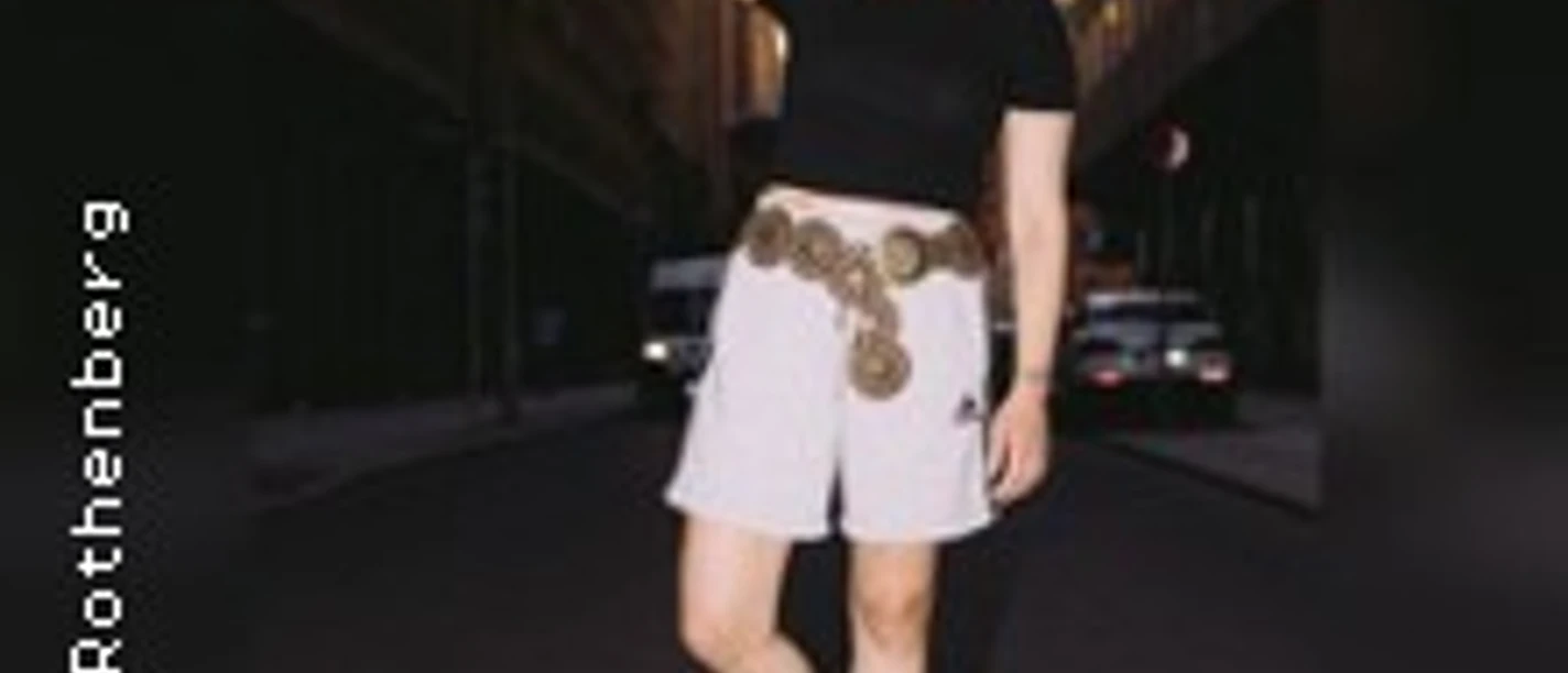 Eine Person mit schwarzem Oberteil und weißen Shorts posiert selbstbewusst in einer urbanen Umgebung.