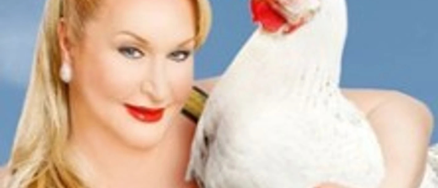 Eine lächelnde Person mit blonden Haaren hält ein weißes Huhn vor einem klaren blauen Himmel.