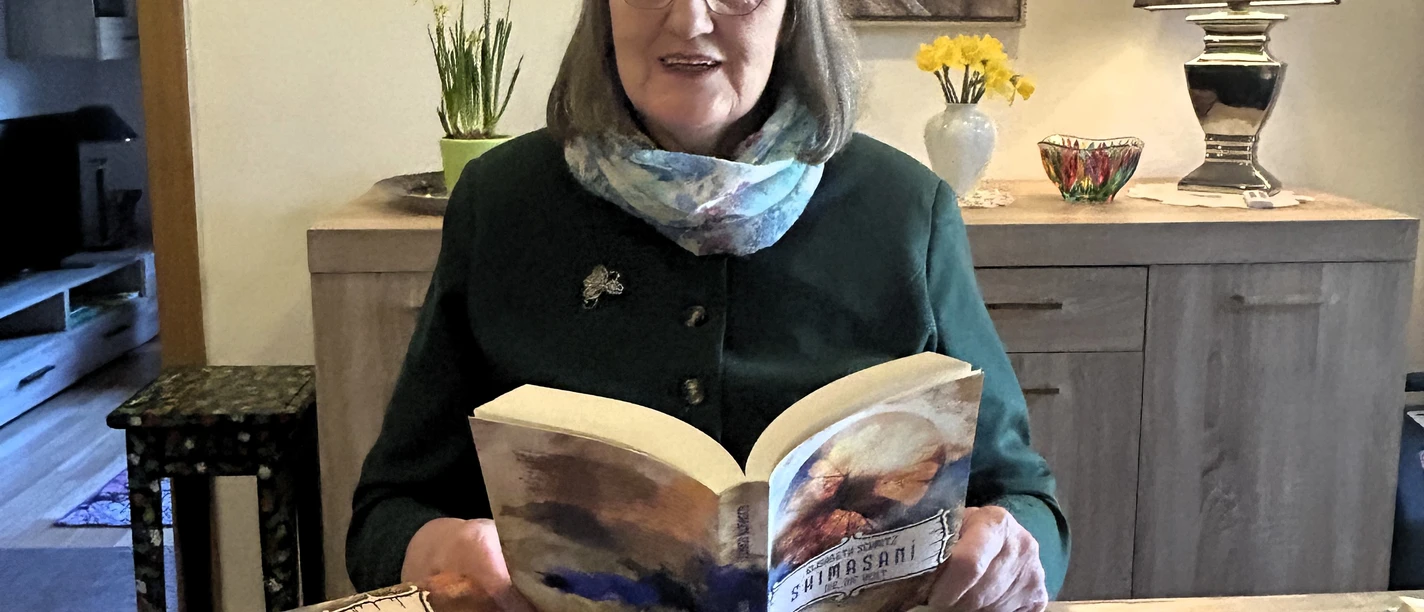 Elisabeth Schmitz_Portrait quer.JPG Frau mit Brille liest an einem Tisch in hellem Raum ein Buch, weitere Bücher liegen vor ihr.