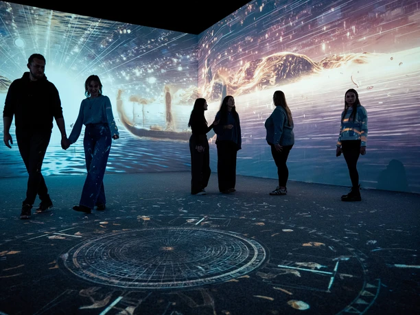 TUTANCHAMUN – Ein immersives Abenteuer Eine Gruppe von Menschen bewegt sich in einem dunklen Raum, umgeben von ägyptischen Lichtprojektionen.