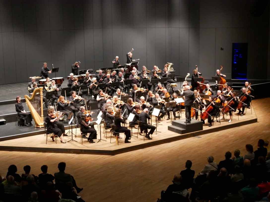 Orchester Osnabrücker Musikfreunde Orchester spielt auf einer Bühne, Musiker in formeller Kleidung konzentrieren sich auf Instrumente.