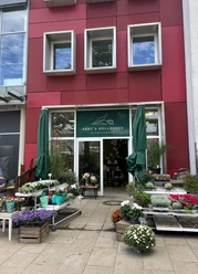 anneshollaender.jpg Außenbereich eines Blumengeschäfts mit roter Fassade. Vor der Eingang stehen rechts links niedrige Regale mit frischen, bunten Pflanzen.