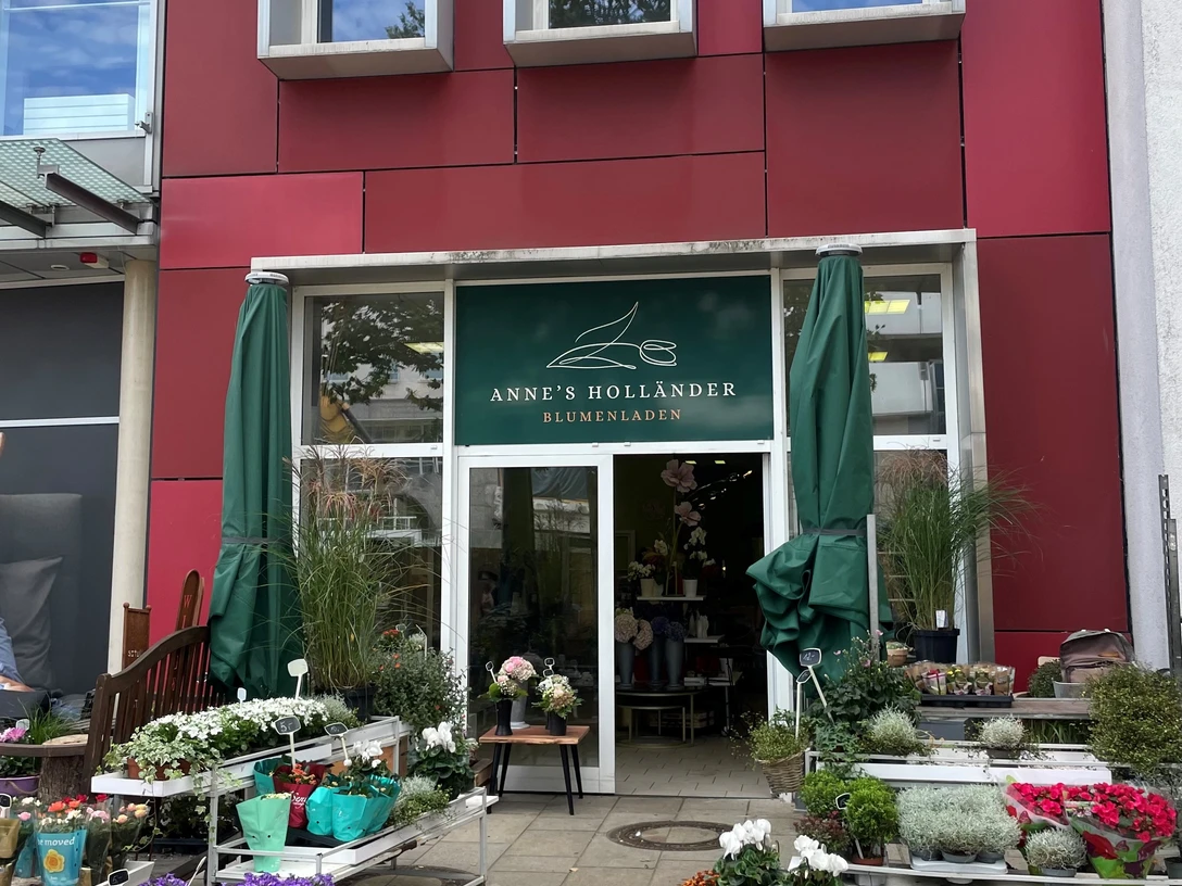 anneshollaender.jpg Außenbereich eines Blumengeschäfts mit roter Fassade. Vor der Eingang stehen rechts links niedrige Regale mit frischen, bunten Pflanzen.