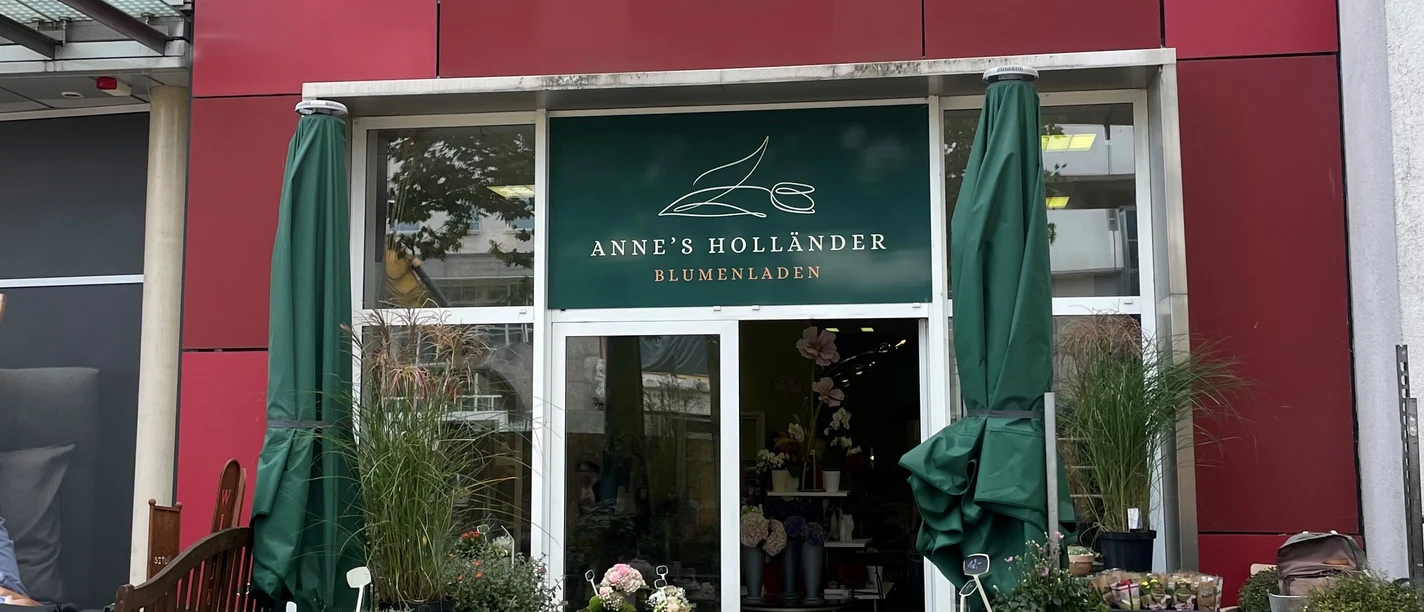 anneshollaender.jpg Außenbereich eines Blumengeschäfts mit roter Fassade. Vor der Eingang stehen rechts links niedrige Regale mit frischen, bunten Pflanzen.