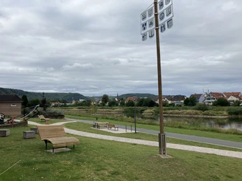 An der Weserpromenade stehen ein Gewerkebaum und moderne Holzliegen und Bänke zum Entspannen, dazwischen verlaufen Wege, im Hintergrund ist der Fluss und der Nachbarort zu sehen.