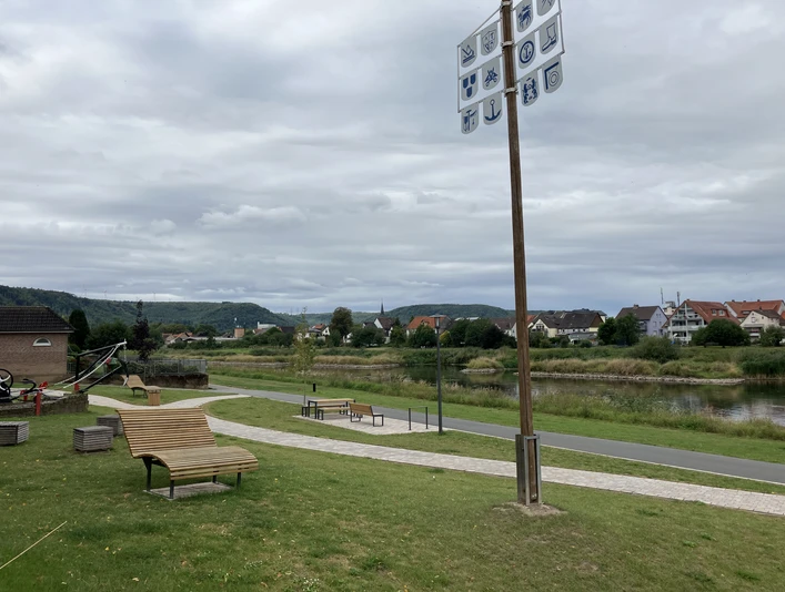 Weserpromenade in Herstelle An der Weserpromenade stehen ein Gewerkebaum und moderne Holzliegen und Bänke zum Entspannen, dazwischen verlaufen Wege, im Hintergrund ist der Fluss und der Nachbarort zu sehen.