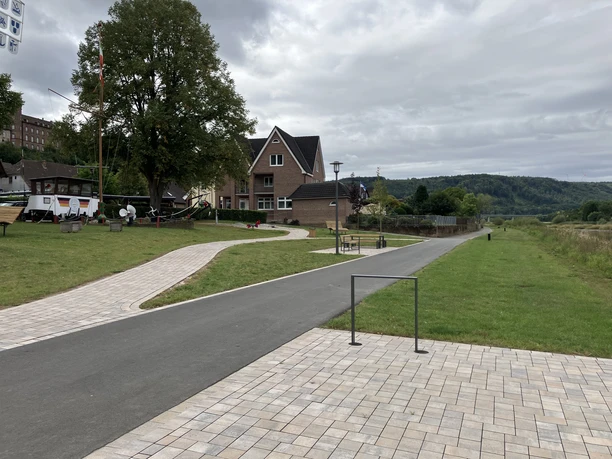 Das Weserufer wird durch Wiesenflächen sowie gepflasterte und asphaltierte Wege gekennzeichnet, ergänzt durch Fahrradbügel und Sitzmöbel. Im Hintergrund links ist ein alter Schiffsrumpf zu sehen.