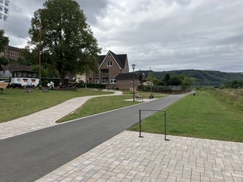 Weserufer in Herstelle Das Weserufer wird durch Wiesenflächen sowie gepflasterte und asphaltierte Wege gekennzeichnet, ergänzt durch Fahrradbügel und Sitzmöbel. Im Hintergrund links ist ein alter Schiffsrumpf zu sehen.