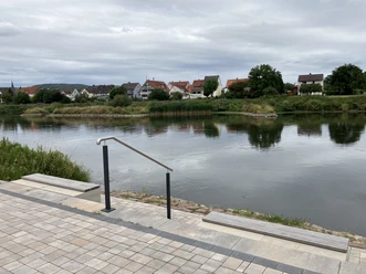Gepflasterte Sitzstufen mit Holzbänken ermöglichen eine Pause direkt am Fluss. Im Hintergrund ist auf der anderen Flussseite der Nachbarort zu sehen.