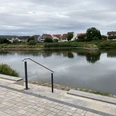 Gepflasterte Sitzstufen mit Holzbänken ermöglichen eine Pause direkt am Fluss. Im Hintergrund ist auf der anderen Flussseite der Nachbarort zu sehen.