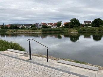 Sitzstufen an der Weserpromenade in Herstelle Gepflasterte Sitzstufen mit Holzbänken ermöglichen eine Pause direkt am Fluss. Im Hintergrund ist auf der anderen Flussseite der Nachbarort zu sehen.