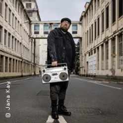 Ein Mensch mit Kappe und Brille steht vor städtischer Kulisse und hält einen großen Ghettoblaster.