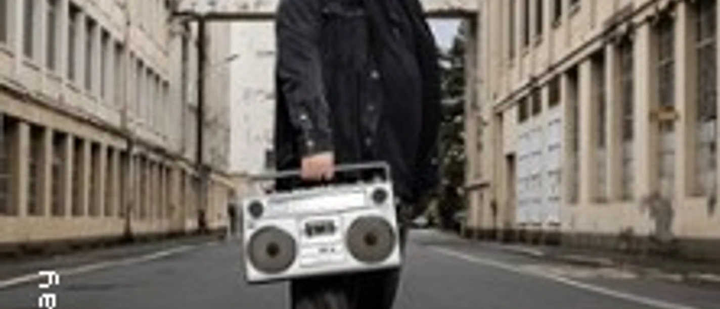 Ein Mensch mit Kappe und Brille steht vor städtischer Kulisse und hält einen großen Ghettoblaster.