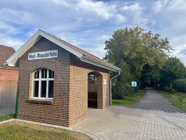 Historische Schutzhütte am Eingang Ostfriesland-Wanderweg Ziegelsteingebäude mit einem Satteldach, beschriftet mit West- Rhauderfehn. Vor dem Gebäude führt ein gepflasterter Weg in einen bewaldeten Wegbereich.