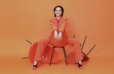 anna-depenbusch-c-steven-haberland-quer-cmyk (1).jpg Frau in orangefarbenem Anzug sitzt lässig auf Stuhl vor orangefarbenem Hintergrund und lacht