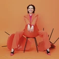 anna-depenbusch-c-steven-haberland-quer-cmyk (1).jpg Frau in orangefarbenem Anzug sitzt lässig auf Stuhl vor orangefarbenem Hintergrund und lacht