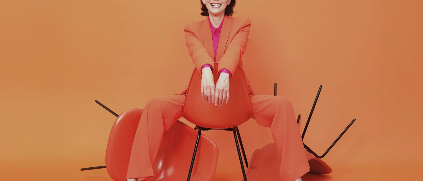 anna-depenbusch-c-steven-haberland-quer-cmyk (1).jpg Frau in orangefarbenem Anzug sitzt lässig auf Stuhl vor orangefarbenem Hintergrund und lacht