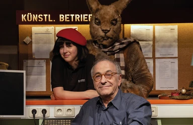 theater-laboratorium-c-theater-laboratorium-quer-cmyk-2.jpg Drei Personen in einem Theaterbüro, darunter eine Person im Känguru-Kostüm, freundlich lächelnd.