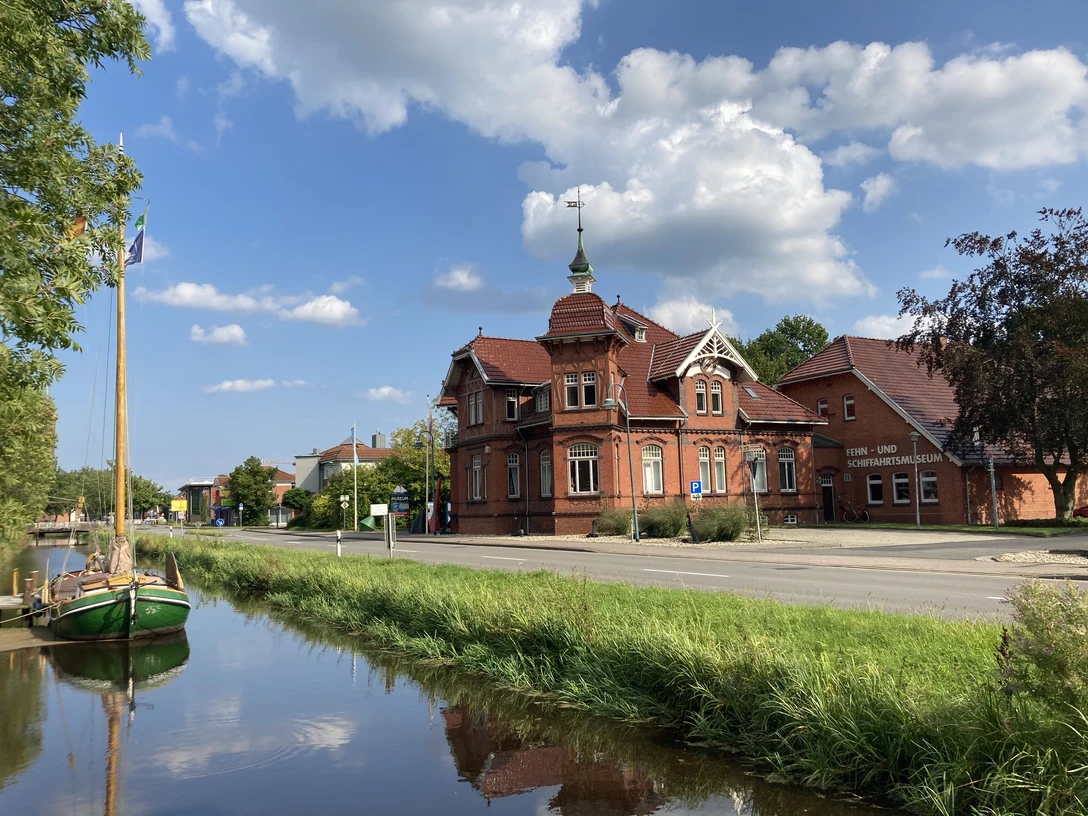 Fehn-und Schiffahrtsmuseum Westrhauderfehn Zwei Gebäude in Backsteinbauweise, eines mit einem Turm und einem Schornstein, am Straßenrand. Im Vordergrund ein schmaler Kanal mit einem Boot.