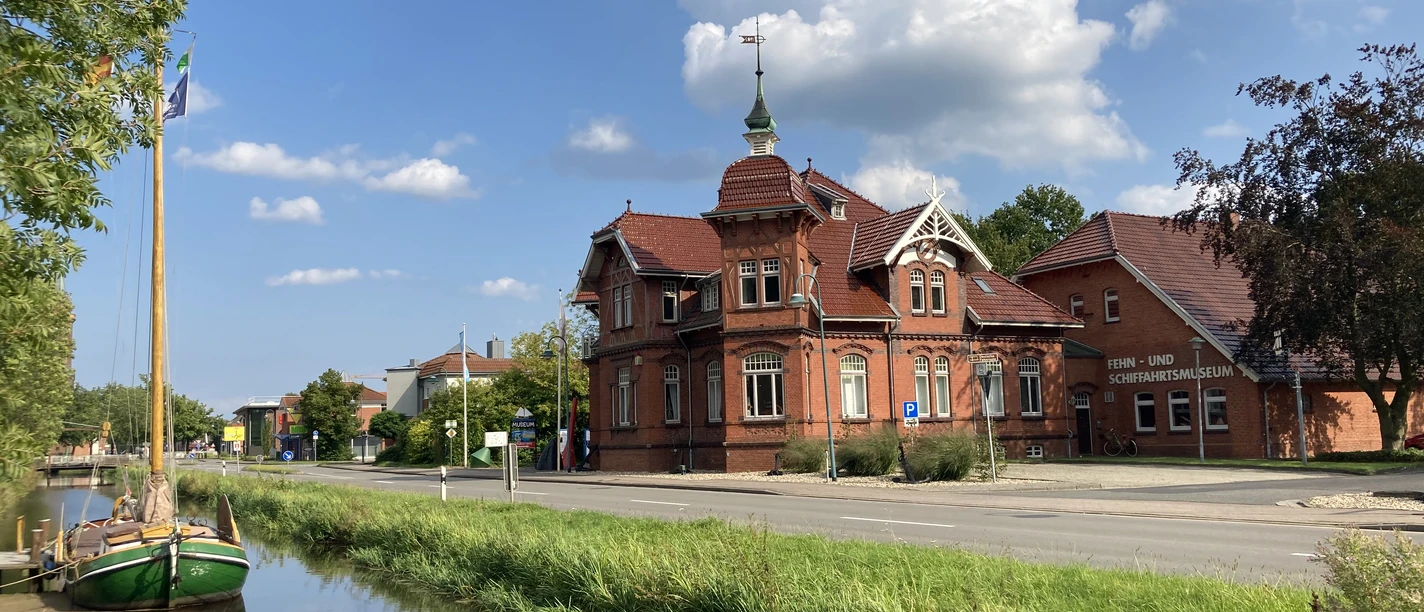 Fehn-und Schiffahrtsmuseum Westrhauderfehn Zwei Gebäude in Backsteinbauweise, eines mit einem Turm und einem Schornstein, am Straßenrand. Im Vordergrund ein schmaler Kanal mit einem Boot.