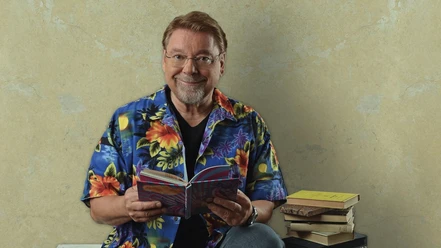 Jurgen-von-der-Lippe-_c_-Andre-Kowalski.png Ein Mann mit Brille in bunter Blumenhemd sitzt lächelnd mit Buch neben einem Stapel weiterer Bücher.