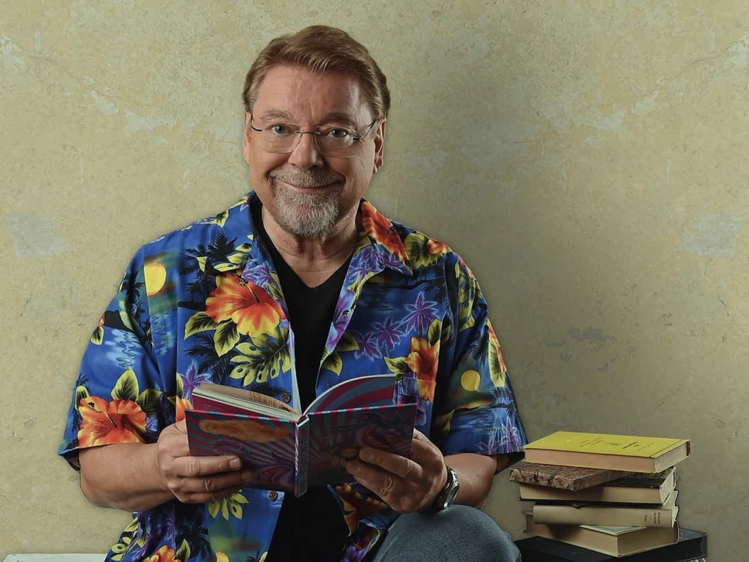 Jurgen-von-der-Lippe-_c_-Andre-Kowalski.png Ein Mann mit Brille in bunter Blumenhemd sitzt lächelnd mit Buch neben einem Stapel weiterer Bücher.