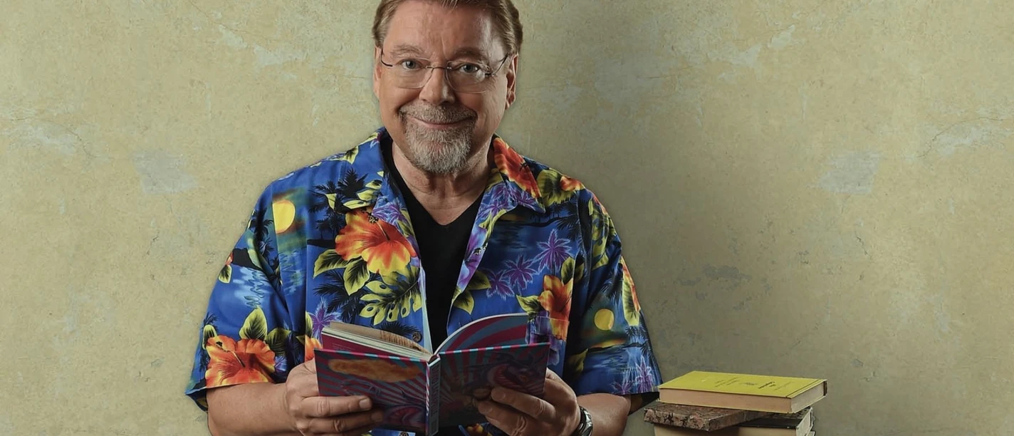 Jurgen-von-der-Lippe-_c_-Andre-Kowalski.png Ein Mann mit Brille in bunter Blumenhemd sitzt lächelnd mit Buch neben einem Stapel weiterer Bücher.