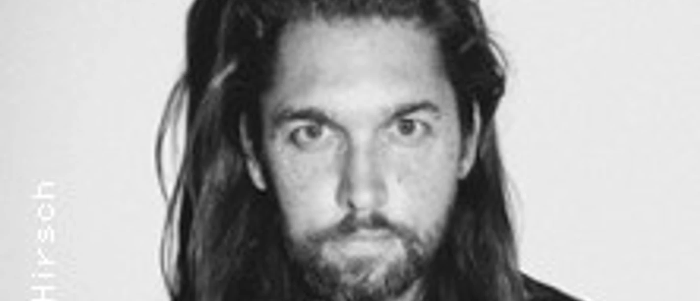 Porträt eines Mannes mit langem Haar und Bart vor neutralem Hintergrund, leicht ernst wirkender Ausdruck.