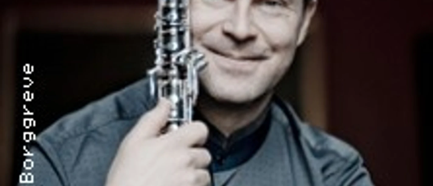 Ein lächelnder Mann hält eine Klarinette und trägt ein elegantes, dunkles, langärmeliges Hemd.