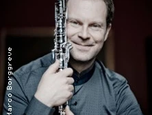 Ein lächelnder Mann hält eine Klarinette und trägt ein elegantes, dunkles, langärmeliges Hemd.