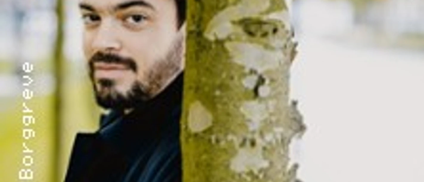 Ein Mann mit dunklem Haar und Bart lehnt an einem Baum, blickt freundlich in die Kamera mit unscharfem Hintergrund.