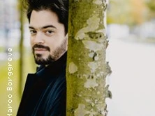 Ein Mann mit dunklem Haar und Bart lehnt an einem Baum, blickt freundlich in die Kamera mit unscharfem Hintergrund.