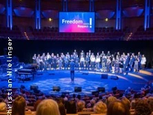 Konzertbühne mit einer großen Chorgruppe im Halbkreis beleuchtet, Publikum im Vordergrund.