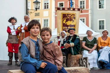 Der-Retter-der-Stadt-Pirna-(C)-Sven_Lehmann_1920x1280px_MG_7659.jpg Zwei Jungen in historischer Kleidung sitzen auf Holzbank, Erwachsene in traditioneller Tracht im Hintergrund.