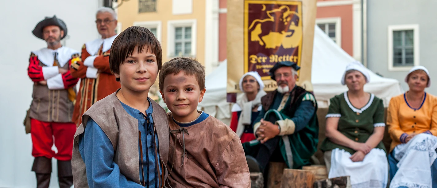 Der-Retter-der-Stadt-Pirna-(C)-Sven_Lehmann_1920x1280px_MG_7659.jpg Zwei Jungen in historischer Kleidung sitzen auf Holzbank, Erwachsene in traditioneller Tracht im Hintergrund.