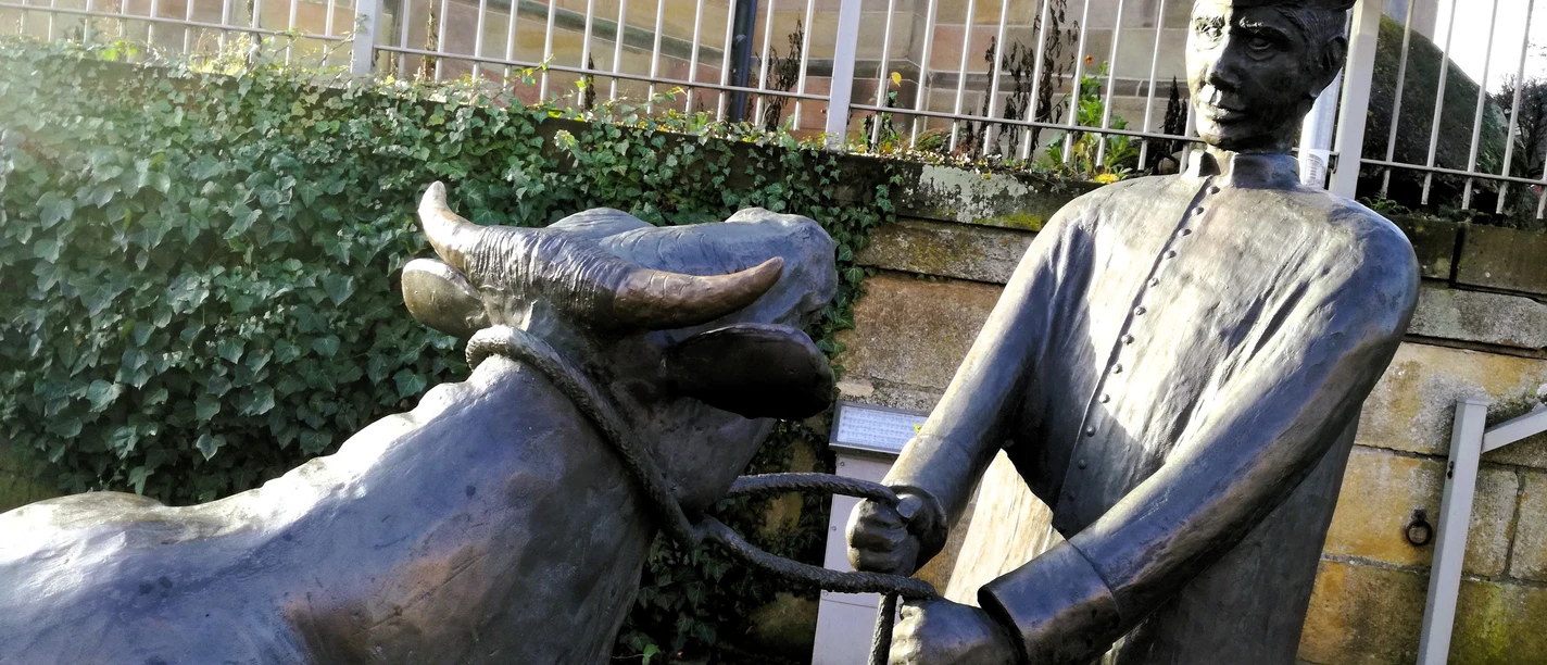 Denkmal Pastor sine Koh in Emsbüren Bronzestatue eines Pastors mit einer Kuh an einem Seil vor einer Mauer mit Wegweisern in Emsbüren.
